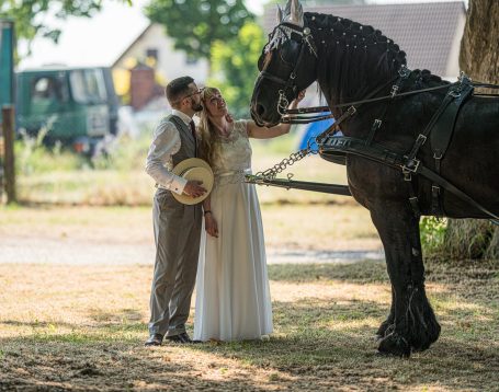 Ein Brautpaar küsst sich neben einem mit Pferd gezogenen Wagen in ländlicher Umgebung.