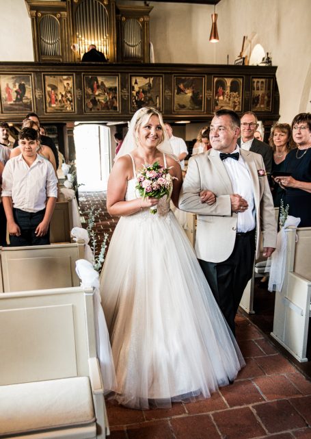 Braut in weißem Kleid mit Blumenstrauß, begleitet von einem Mann, Gäste im Hintergrund.