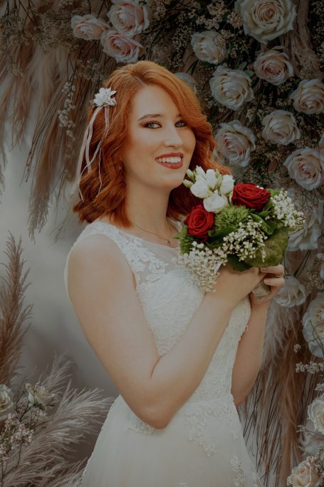 Braut mit rotem Haar, im Hochzeitskleid, lächelt und hält einen Blumenstrauß.