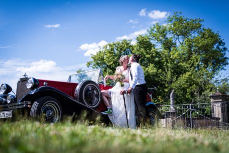 Hochzeitspaar posiert lächelnd neben einem klassischen roten Auto im Freien.