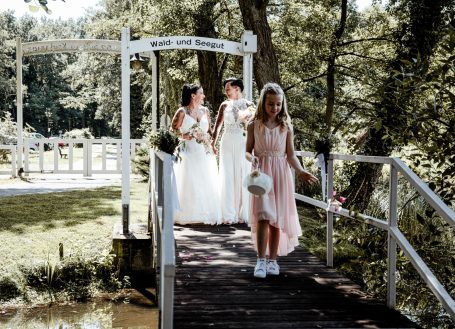 Brautpaar auf einer Brücke, umgeben von Natur, mit einem Mädchen in einem rosa Kleid.