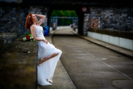 Braut in weißem Kleid, an einer Mauer stehend, mit roten Blumen und umgeben von Natur.