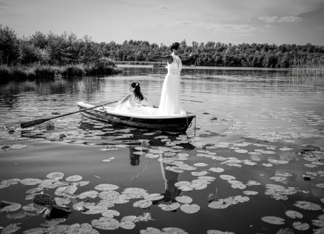 Zwei Frauen in Hochzeitskleidern rudern in einem Boot auf einem See mit Seerosen.