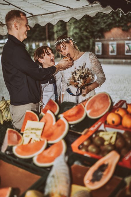 Ein Verkäufer füttert einen Jungen, während eine Frau mit Obstständen posiert.