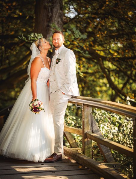 Hochzeitspaar posiert glücklich auf einer Holzbrücke in einer grünen Umgebung.