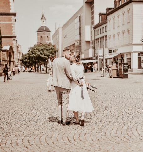 Paar in eleganter Kleidung auf einem Kopfsteinpflasterplatz vor historischer Kulisse.