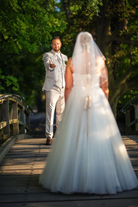 Braut in weißem Kleid auf einer Brücke, Bräutigam in grauem Anzug hält die Hand aus.